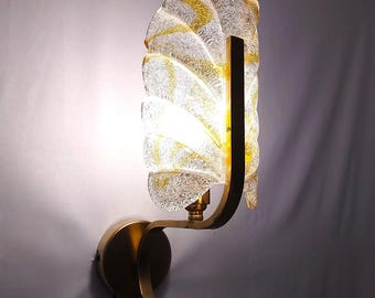 May include: A gold wall sconce with a clear glass shade that has a textured, rippled pattern. The shade is shaped like a leaf and has a gold metal frame.