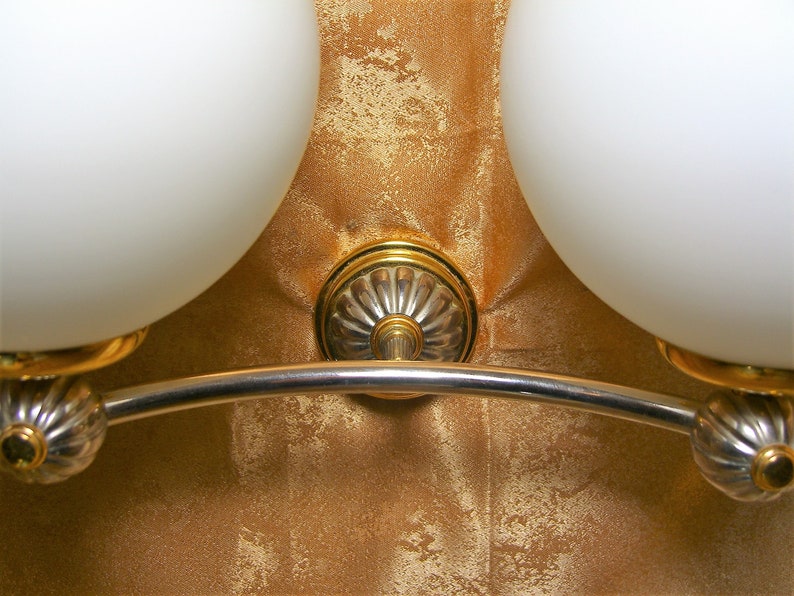 May include: A silver and gold wall-mounted light fixture with two white glass globes. The fixture has a curved arm and decorative details.