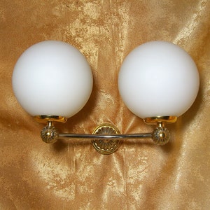 May include: A gold wall sconce with three white globe light fixtures. The sconce has a decorative design with a curved bar connecting the fixtures.