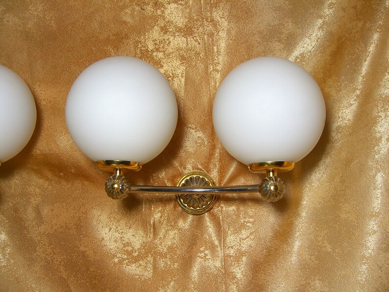 May include: A gold wall sconce with three white globe light fixtures. The sconce has a decorative design with a curved bar connecting the fixtures.