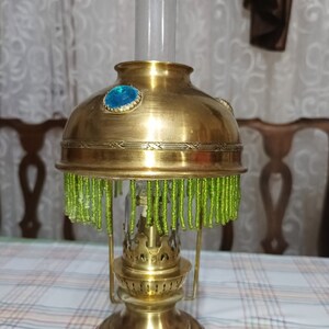 Op de afbeelding: Een messing olielamp met een groene kralenrand en een blauwe glazen edelsteen aan de bovenkant. De lamp heeft een glazen schoorsteen en een decoratieve basis.