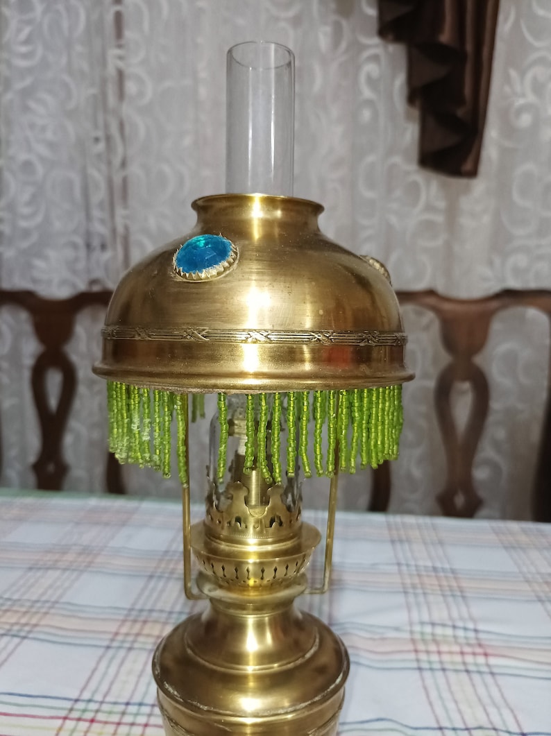 Op de afbeelding: Een messing olielamp met een groene kralenrand en een blauwe glazen edelsteen aan de bovenkant. De lamp heeft een glazen schoorsteen en een decoratieve basis.