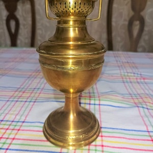 Op de afbeelding: Een gouden messing olielamp met een decoratief, versierd ontwerp. De lamp heeft een ronde basis en een cilindrisch lichaam met een decoratieve bovenkant.