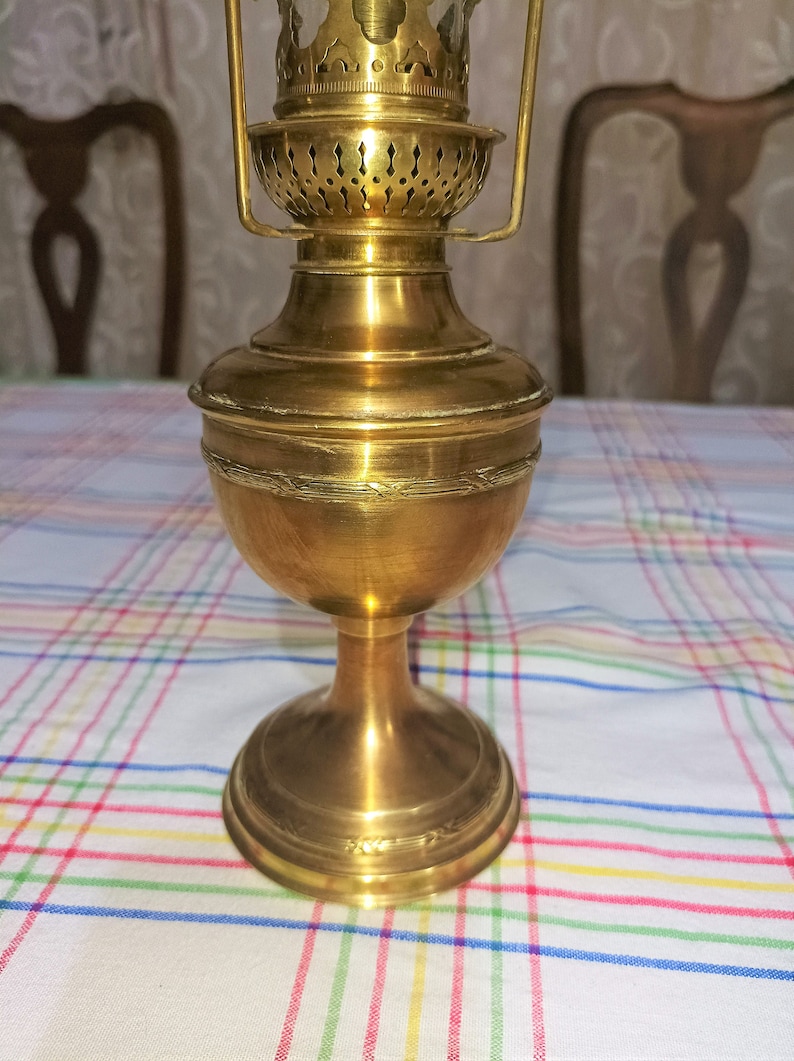 Op de afbeelding: Een gouden messing olielamp met een decoratief, versierd ontwerp. De lamp heeft een ronde basis en een cilindrisch lichaam met een decoratieve bovenkant.