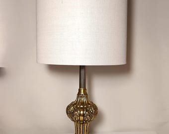May include: A vintage table lamp with a gold-colored, ornate base and a cylindrical white lampshade. The lamp features a detailed, decorative design on the base and stem. The lampshade is made of a light-colored fabric.