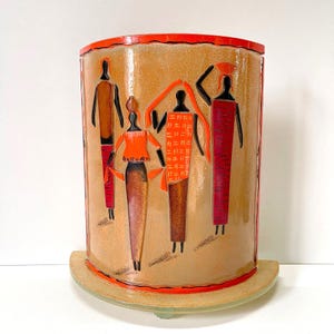 Puede incluir: Una base de lámpara cilíndrica de cerámica con fondo beige y un diseño colorido de cuatro figuras estilizadas en naranja, rojo y marrón. Las figuras visten ropa tradicional y están dispuestas en línea. La base de la lámpara tiene un fondo redondeado y un borde rojo.