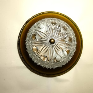 Può includere: Un lampadario vintage con un paralume in vetro trasparente a motivi e una base in metallo color ottone. Il paralume in vetro ha un design floreale e un centro in ottone. L'apparecchio è montato su un soffitto bianco.