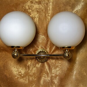 May include: A gold wall sconce with two white frosted glass globes. The sconce has a decorative design on the arms and a center piece.