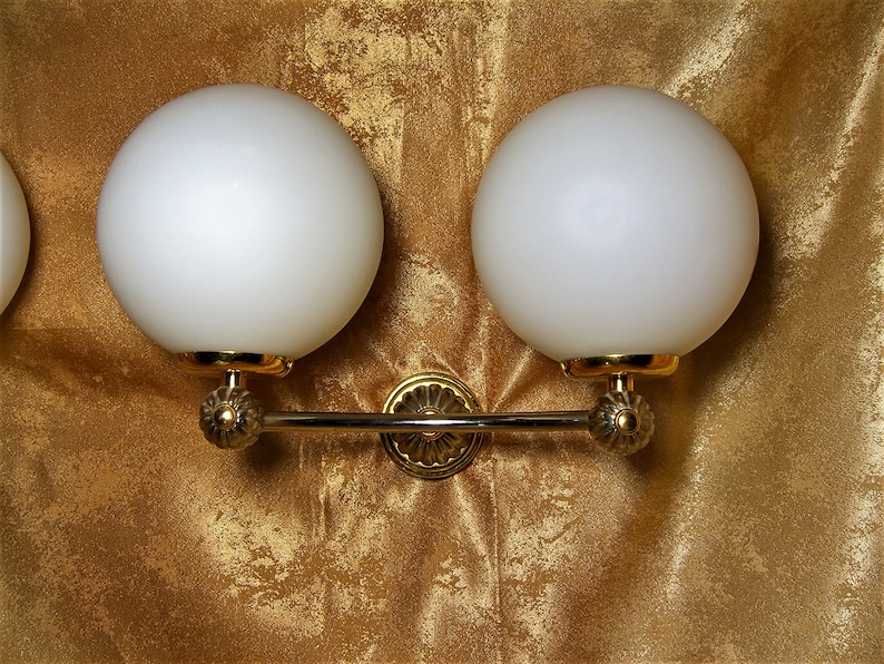 May include: A gold wall sconce with two white frosted glass globes. The sconce has a decorative design on the arms and a center piece.