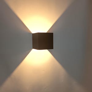 May include: A square, dark-coloured wall-mounted light fitting. The light shines upwards and downwards, creating a fan-like pattern on the wall. The light is a warm, yellow colour.
