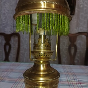 Op de afbeelding: Een messing olielamp met een groene parelkap en een blauwe glazen accent. De lamp heeft een decoratief ontwerp en een glazen schoorsteen.