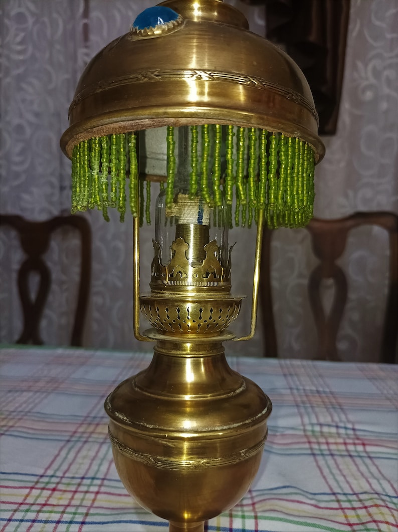 Op de afbeelding: Een messing olielamp met een groene parelkap en een blauwe glazen accent. De lamp heeft een decoratief ontwerp en een glazen schoorsteen.