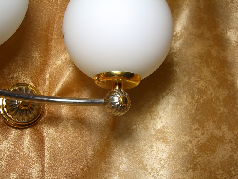 May include: A silver and gold wall sconce with two white glass globes. The sconce has a curved arm and decorative details.