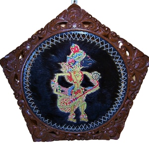 Puede incluir: Un marco de madera circular con tallas intrincadas, que presenta un fondo de cuero negro con una figura pintada de colores en el centro. La figura es una marioneta de sombras tradicional indonesia, conocida como wayang kulit, con detalles intrincados y colores vibrantes.