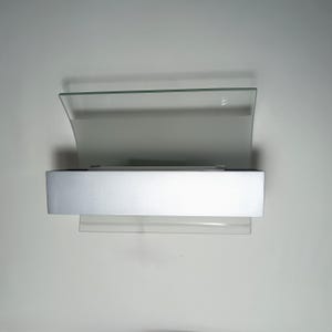 May include: A modern wall sconce with a frosted glass shade and a silver-colored metal base. The sconce has a rectangular shape with a curved top and a clear glass shelf below.
