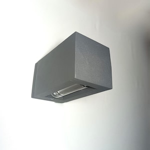 May include: A gray, rectangular outdoor wall light fixture. The fixture has a boxy design with a recessed light source. The light is mounted on a white wall. The fixture is approximately 6 inches wide and 4 inches tall.