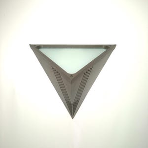May include: A triangular, modern wall sconce with a frosted glass panel. The sconce has a metallic, possibly aluminum, frame with a series of angled lines on the sides. The light fixture is designed to be mounted on a wall and provides ambient lighting.