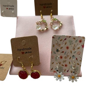 Könnte beinhalten: Eine Auswahl handgefertigter Ohrringe, präsentiert auf rosa Stoff. Die Ohrringe umfassen goldene Creolen, Blumenkränze, rote Edelstein-Tropfen und Daisy-Ohrringe. Jedes Paar ist an einer braunen Karte mit dem Text "Handmade with love" befestigt.