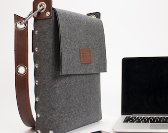 May include: A gray felt laptop bag with a brown leather strap and silver rivets. The bag has a flap closure and a small leather patch on the front. The bag is sitting on a white surface next to a laptop, a smartphone, and a red notebook.