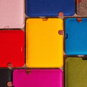 May include: A selection of brightly coloured tablet sleeves made of felt. The sleeves are in shades of red, yellow, blue, and green. Each has a rectangular shape, a small cutout, and a copper-coloured clasp. The sleeves are arranged closely together.