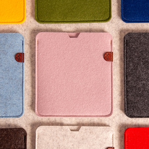 Puede incluir: Una colección de fundas rectangulares de fieltro en varios colores, incluyendo amarillo, verde oliva, azul, azul claro, rosa, gris oscuro, marrón, blanco roto y rojo. Cada funda presenta una pequeña lengüeta de cuero marrón en el lateral.