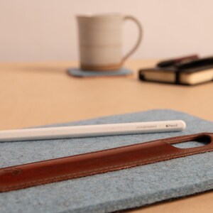 Puede incluir: Un l&aacute;piz &oacute;ptico blanco y un estuche de cuero marr&oacute;n para bol&iacute;grafo descansan sobre una superficie de fieltro azul claro. El l&aacute;piz &oacute;ptico tiene el texto "JACK&JONES" y "Pencil". Una taza y un cuaderno est&aacute;n en el fondo.