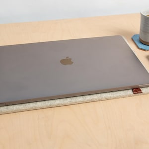 May include: A silver laptop with the Apple logo sits on a light grey felt laptop sleeve. A white mug with tea is on a blue coaster, and a black smartphone is nearby. The items are on a light wood surface.
