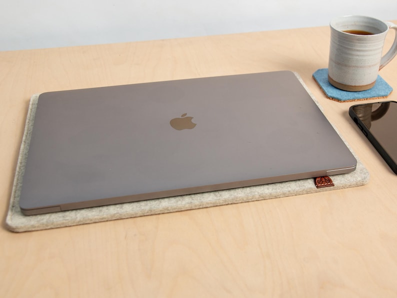 May include: A silver laptop with the Apple logo sits on a light grey felt laptop sleeve. A white mug with tea is on a blue coaster, and a black smartphone is nearby. The items are on a light wood surface.