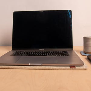 May include: A silver laptop with a black screen sits on a beige felt pad. A ceramic mug rests on a blue coaster, and a smartphone is placed to the right. The items are on a light wood surface.