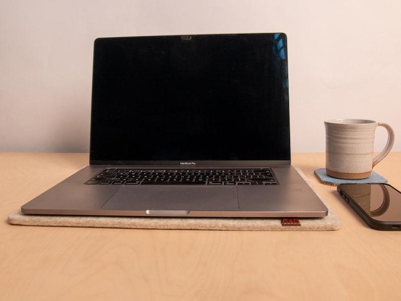 May include: A silver laptop with a black screen sits on a beige felt pad. A ceramic mug rests on a blue coaster, and a smartphone is placed to the right. The items are on a light wood surface.