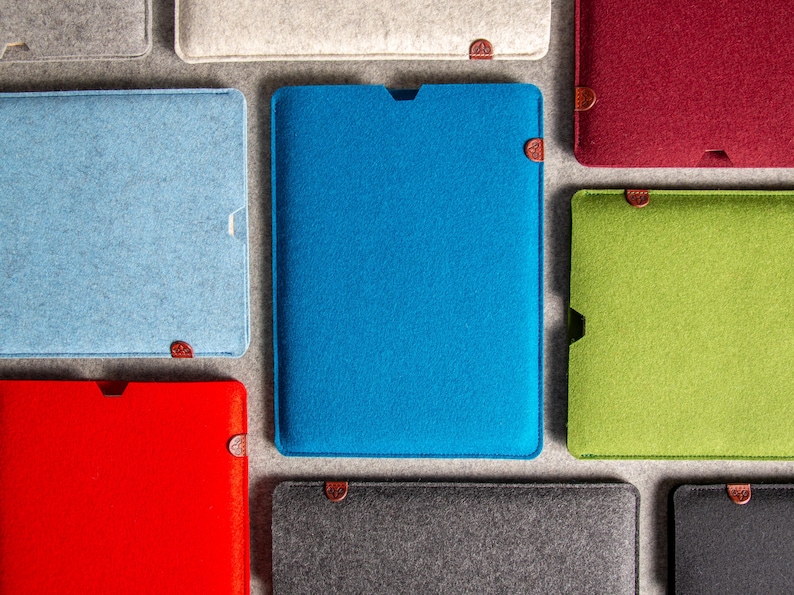May include: A selection of colourful felt tablet sleeves in shades of blue, red, and green. Each sleeve is rectangular with a curved top and a small, circular fastener. The sleeves are arranged on a neutral grey surface.