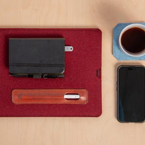 Puede incluir: Un organizador de escritorio de fieltro burdeos con un cuaderno negro, un bol&iacute;grafo y un portal&aacute;pices de cuero. Una taza blanca con caf&eacute; se encuentra sobre un posavasos azul, junto a un tel&eacute;fono inteligente. Los art&iacute;culos est&aacute;n dispuestos sobre una superficie de madera clara.