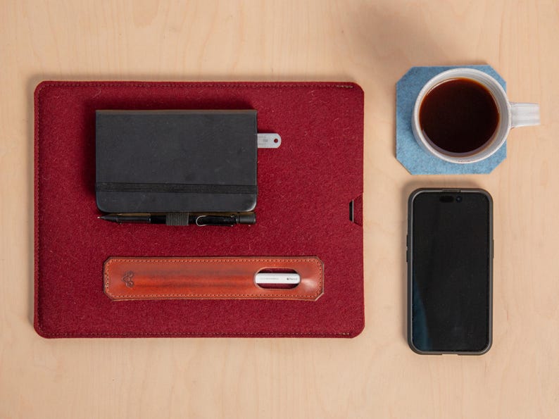 Puede incluir: Un organizador de escritorio de fieltro burdeos con un cuaderno negro, un bol&iacute;grafo y un portal&aacute;pices de cuero. Una taza blanca con caf&eacute; se encuentra sobre un posavasos azul, junto a un tel&eacute;fono inteligente. Los art&iacute;culos est&aacute;n dispuestos sobre una superficie de madera clara.
