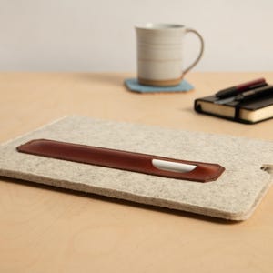 Puede incluir: Una funda para port&aacute;til de fieltro gris claro con un portal&aacute;pices de cuero marr&oacute;n. Un l&aacute;piz &oacute;ptico blanco est&aacute; en el portal&aacute;pices. Una taza, un posavasos y un cuaderno con bol&iacute;grafos est&aacute;n en el fondo.