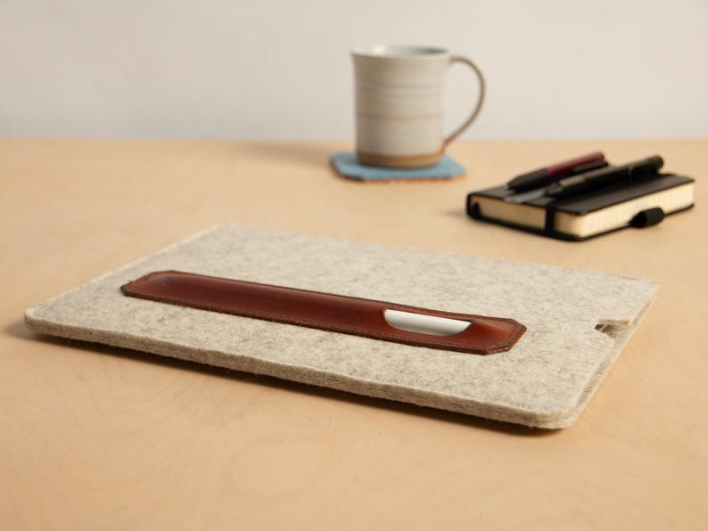 Puede incluir: Una funda para port&aacute;til de fieltro gris claro con un portal&aacute;pices de cuero marr&oacute;n. Un l&aacute;piz &oacute;ptico blanco est&aacute; en el portal&aacute;pices. Una taza, un posavasos y un cuaderno con bol&iacute;grafos est&aacute;n en el fondo.