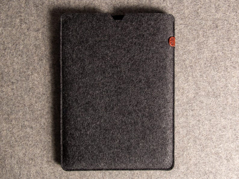 May include: A dark grey felt tablet sleeve with a small, rounded, reddish-brown accent on the right side. The sleeve is rectangular with a slightly curved top edge. The background is a lighter grey felt.