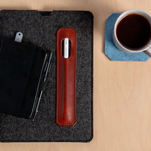 Puede incluir: Vista a&eacute;rea de un espacio de trabajo con un cuaderno negro, un bol&iacute;grafo, un estuche de cuero marr&oacute;n para l&aacute;piz &oacute;ptico y un l&aacute;piz &oacute;ptico blanco. Una taza de caf&eacute; se encuentra sobre un posavasos de fieltro azul, todo sobre una superficie de madera clara.