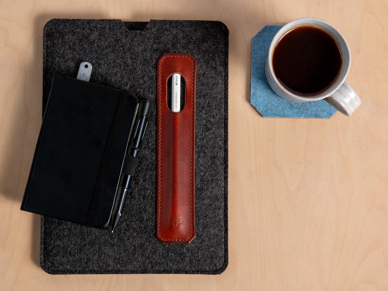 Puede incluir: Vista a&eacute;rea de un espacio de trabajo con un cuaderno negro, un bol&iacute;grafo, un estuche de cuero marr&oacute;n para l&aacute;piz &oacute;ptico y un l&aacute;piz &oacute;ptico blanco. Una taza de caf&eacute; se encuentra sobre un posavasos de fieltro azul, todo sobre una superficie de madera clara.