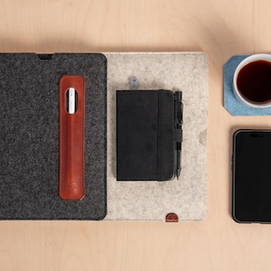 May include: A workspace arrangement seen from above, including a dark grey tablet case, a brown leather stylus holder, a black notebook with a pen, a smartphone, and a coffee mug on a blue coaster. The items are on a light grey and beige felt mat.