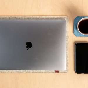 May include: A workspace setup seen from above, including a silver laptop with a black Apple logo, inside a grey felt sleeve. A black smartphone and a coffee mug on a blue coaster are on a light wood surface.