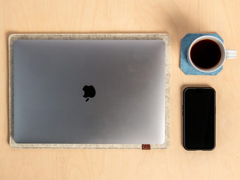 May include: A workspace setup seen from above, including a silver laptop with a black Apple logo, inside a grey felt sleeve. A black smartphone and a coffee mug on a blue coaster are on a light wood surface.