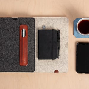 Puede incluir: Vista a&eacute;rea de un espacio de trabajo con una funda de tableta gris oscuro, un soporte para l&aacute;piz t&aacute;ctil de cuero marr&oacute;n, un cuaderno negro con un bol&iacute;grafo, un tel&eacute;fono inteligente y una taza de caf&eacute; sobre un posavasos azul. Los art&iacute;culos est&aacute;n sobre una alfombrilla de fieltro gris claro y beige.
