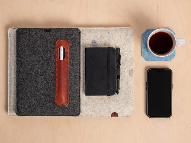 Puede incluir: Vista a&eacute;rea de un espacio de trabajo con una funda de tableta gris oscuro, un soporte para l&aacute;piz t&aacute;ctil de cuero marr&oacute;n, un cuaderno negro con un bol&iacute;grafo, un tel&eacute;fono inteligente y una taza de caf&eacute; sobre un posavasos azul. Los art&iacute;culos est&aacute;n sobre una alfombrilla de fieltro gris claro y beige.