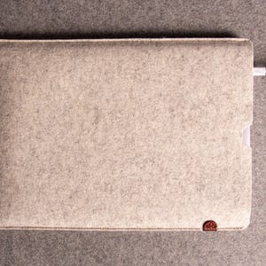 May include: A light grey felt laptop sleeve with a white border and a small brown leather accent. A white charging cable is plugged into the sleeve. The sleeve is rectangular and has a soft, textured appearance.