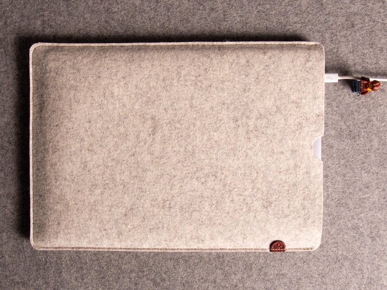 May include: A light grey felt laptop sleeve with a white border and a small brown leather accent. A white charging cable is plugged into the sleeve. The sleeve is rectangular and has a soft, textured appearance.