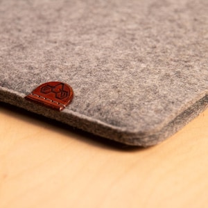 May include: Close-up of a grey felt sleeve with a brown leather patch. The patch features a stitched logo. The sleeve is made of a soft, textured material. The product is likely designed to protect a tablet or laptop.