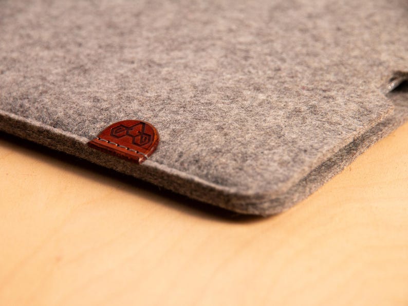 May include: Close-up of a grey felt sleeve with a brown leather patch. The patch features a stitched logo. The sleeve is made of a soft, textured material. The product is likely designed to protect a tablet or laptop.