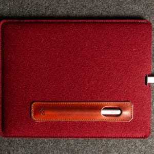 Puede incluir: Una funda de tableta de fieltro burdeos con un acento de cuero marr&oacute;n. El acento de cuero presenta un recorte para un l&aacute;piz &oacute;ptico y un peque&ntilde;o dise&ntilde;o en relieve. Un cable de carga blanco est&aacute; enchufado en la funda.