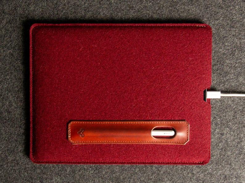 Puede incluir: Una funda de tableta de fieltro burdeos con un acento de cuero marr&oacute;n. El acento de cuero presenta un recorte para un l&aacute;piz &oacute;ptico y un peque&ntilde;o dise&ntilde;o en relieve. Un cable de carga blanco est&aacute; enchufado en la funda.