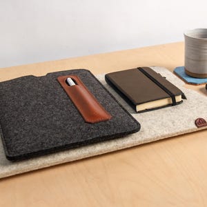 Puede incluir: Una funda de fieltro para port&aacute;til con un portal&aacute;pices de cuero, un cuaderno marr&oacute;n y una taza de cer&aacute;mica sobre una superficie de madera. Tambi&eacute;n hay un smartphone y un posavasos azul. La funda es de color gris oscuro y el cuaderno tiene un cierre el&aacute;stico negro.